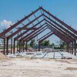 Steel structure of new industrial building under cloud blue sky. New technology structural frame beam of factory in construction. Steel frame manufacturer and pile of sand and gravel in Crosby, TX, US